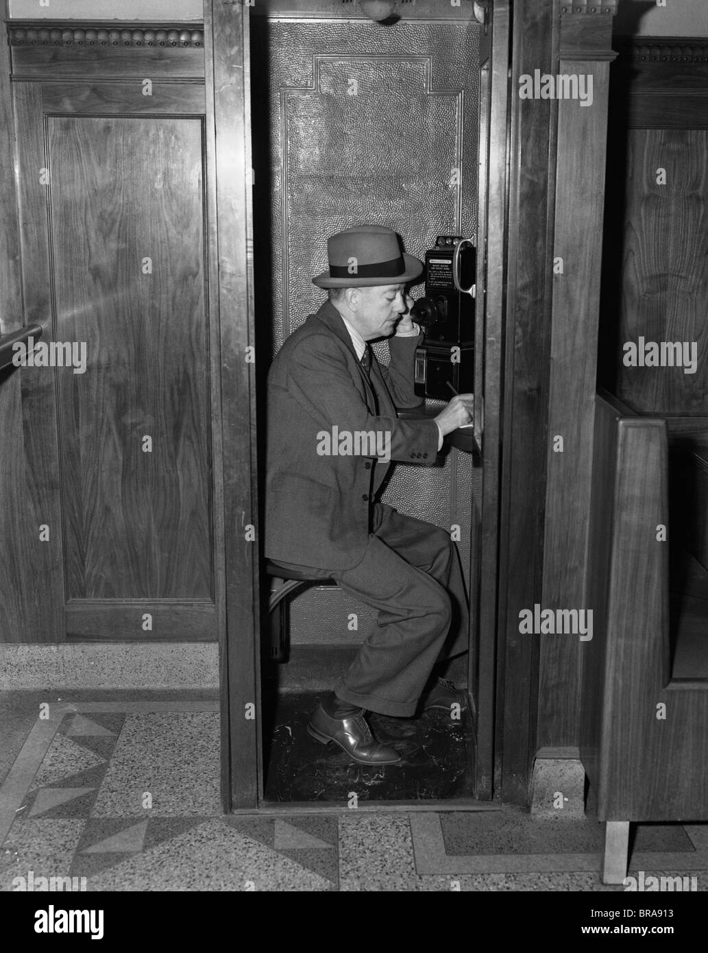 1940ER JAHRE MANN SITZT IN HÖLZERNEN TELEFONZELLE IN EMPFÄNGER SPRECHEN Stockfoto