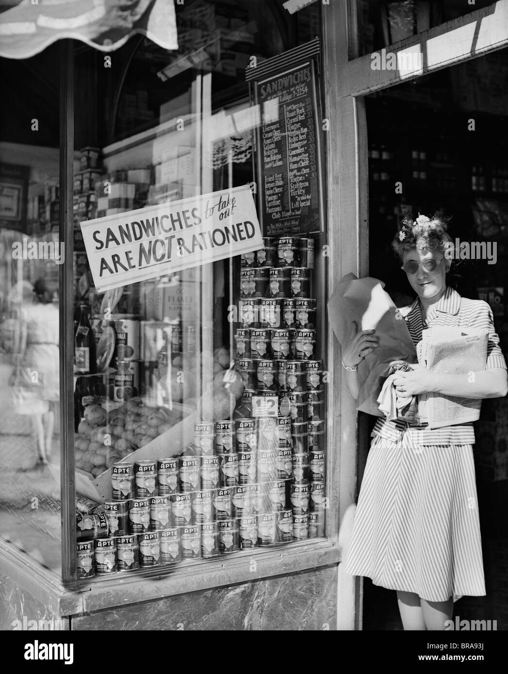 1940ER JAHREN FRAU, DIE PAKETE IN DER TÜR DES NEW YORK CITY DELIKATESSEN MIT EINEM ZEICHEN SANDWICHES ZUM MITNEHMEN SIND NICHT RATIONIERT Stockfoto