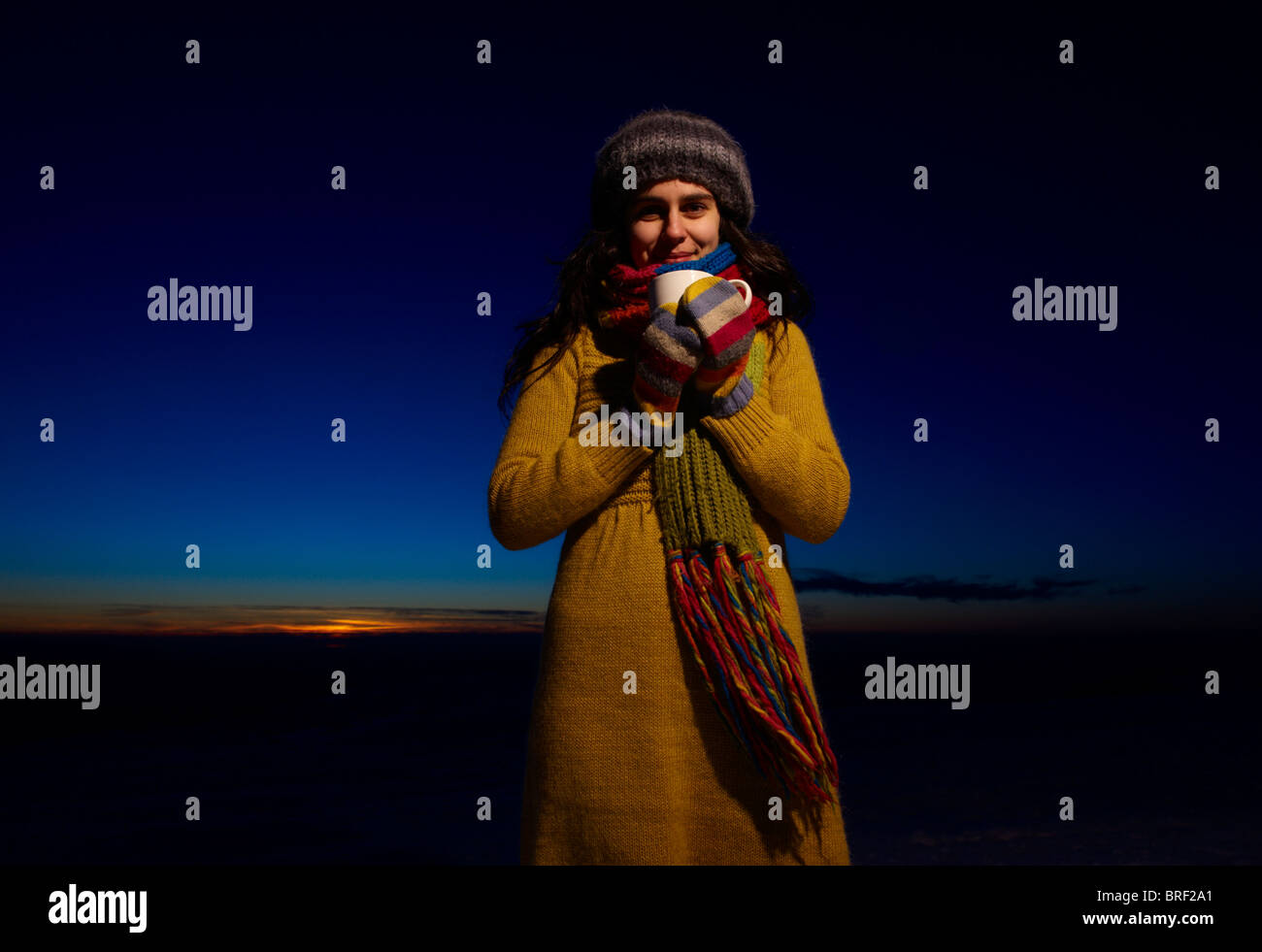 Eine lächelnde junge Frau hält eine Tasse heiße Schokolade vor einem Sonnenuntergang Himmel Stockfoto