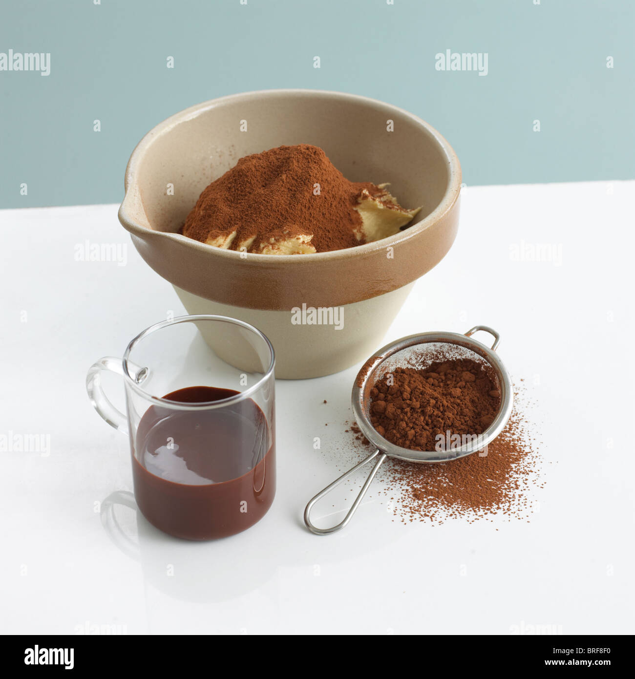 Pudding-Schüssel mit Butter und Kakao Pulver neben Glas geschmolzene Schokolade und Sieb von Schokolade Pulver Stockfoto