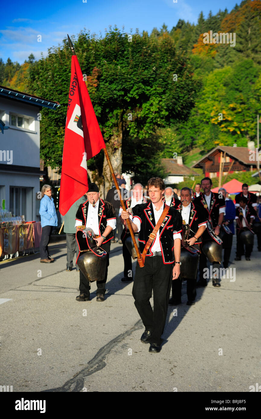 Ein Team von Schweizer Kuh-Glöckner marschieren durch die Stadt St. Cergue, angeführt von einem Mann mit Schweizer Flagge Stockfoto