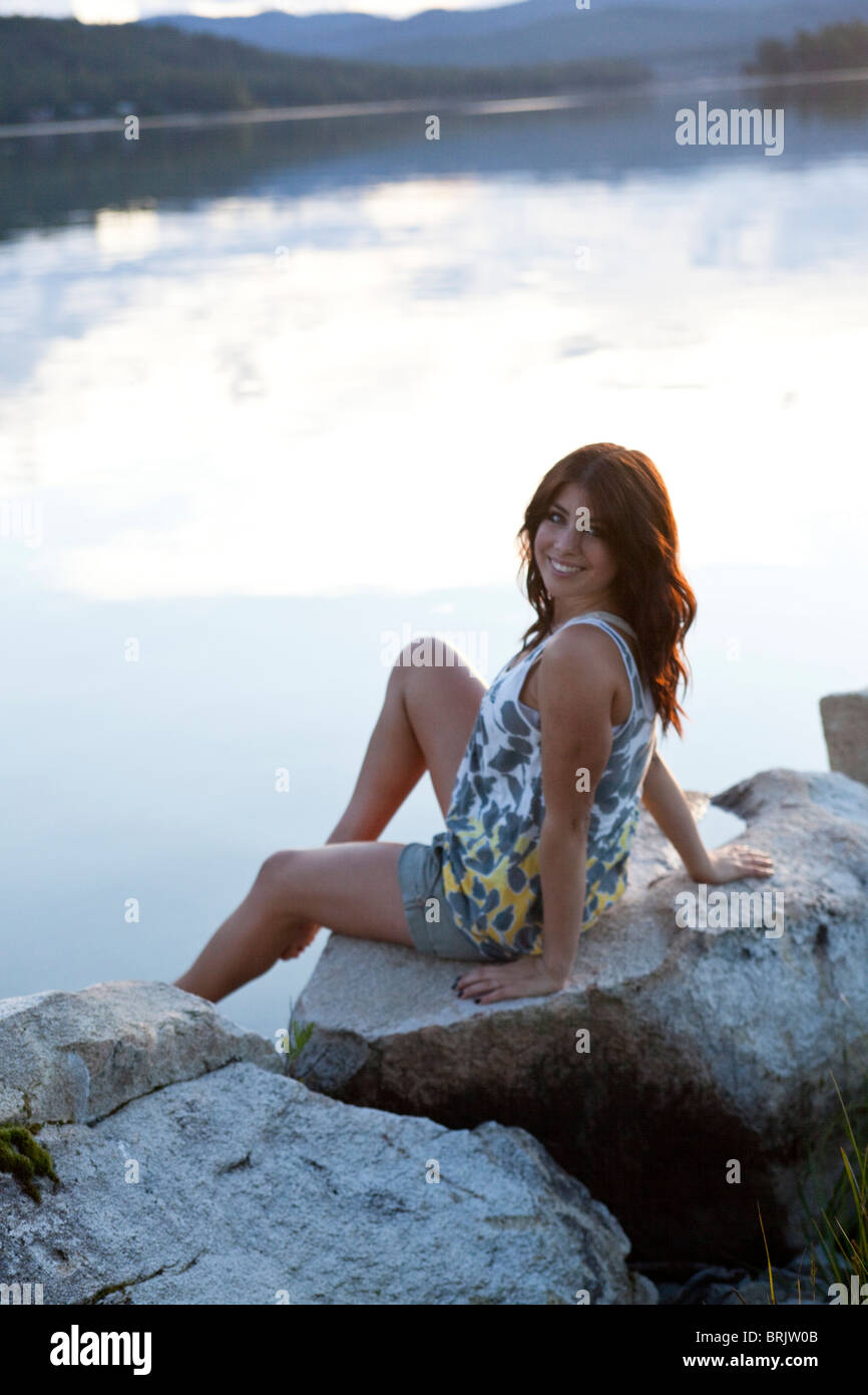 Junge Frau lächelt beim sitzen neben dem See bei Sonnenuntergang. Stockfoto