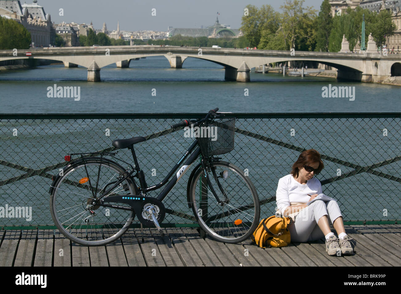 Eine Frau sitzt auf der Pont des Arts neben ihrem Fahrrad, mit der Seine und Pont du Carrousel im Süden in Paris, Frankreich. Stockfoto