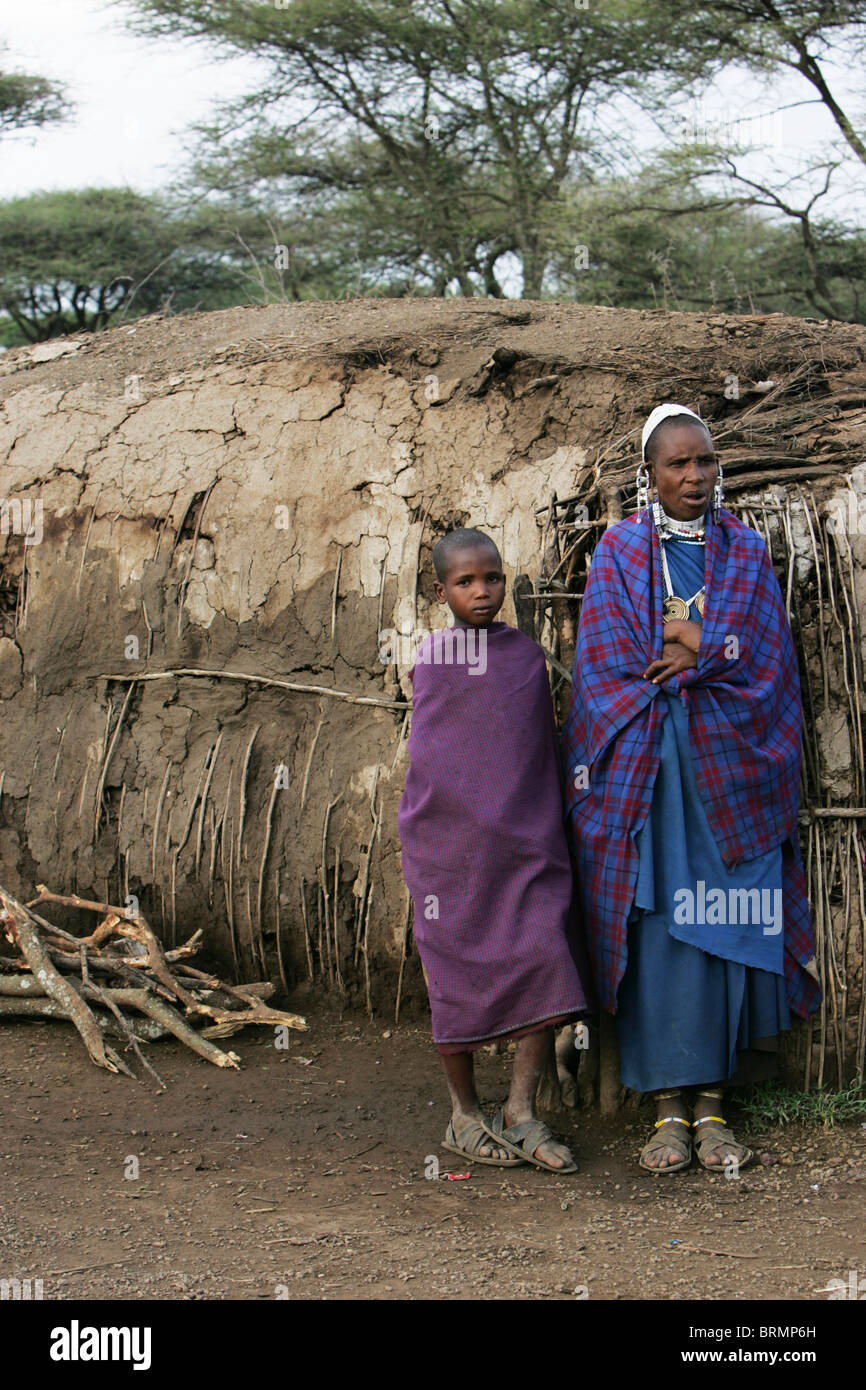 Maasai-Mutter und Kind im Alter von etwa l0 Jahr alt tragen Shukas stehen neben ihren Lehmhütte Stockfoto