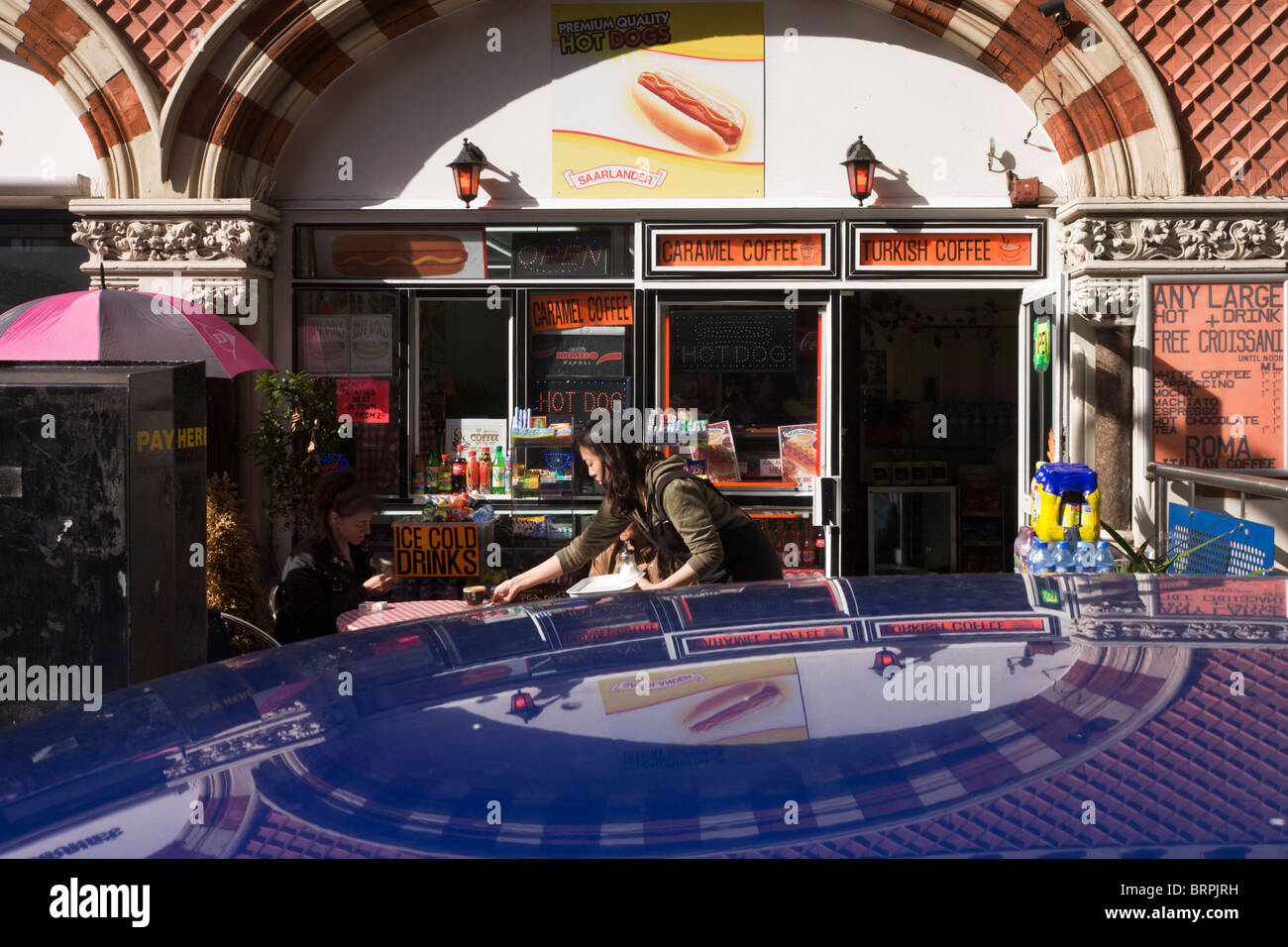 Eine Kellnerin bedient zwei Frauen in einem Straßencafé Straße, unter Edwardian Bögen Architektur und im Autodach gegenüberliegenden reflektiert. Stockfoto