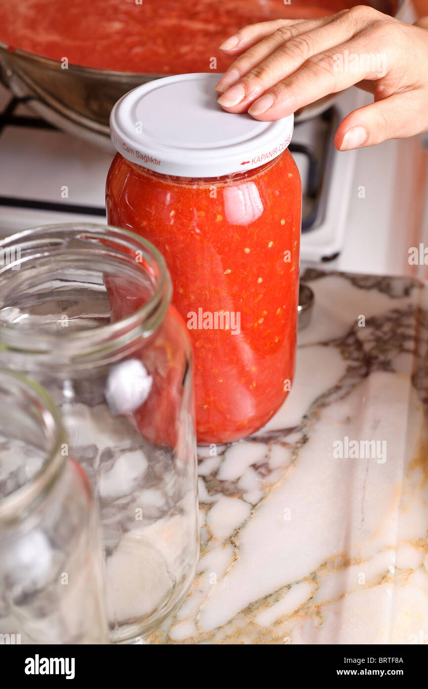 Die Hand einer Frau prüft die Vakuumdichtung eines Deckels auf Glas, gefüllt mit Tomatensaft. Ein leeres Glas ist in der Nähe auf einer Arbeitsplatte Küche Stockfoto