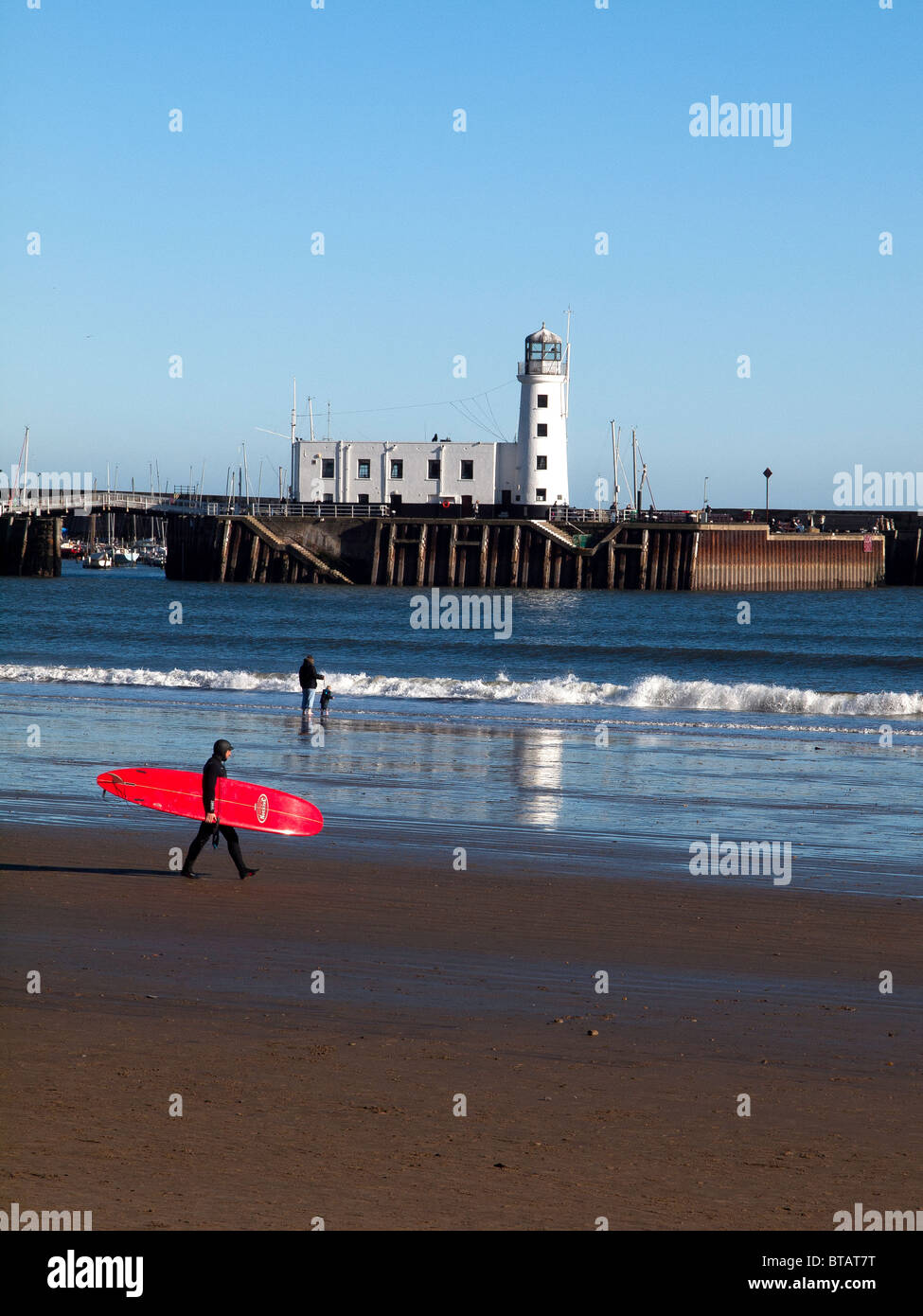 Eine Surfer mit einem leuchtend roten Surfbrett geht über Scarborough South Bay Strand mit dem Leuchtturm im Hintergrund Stockfoto