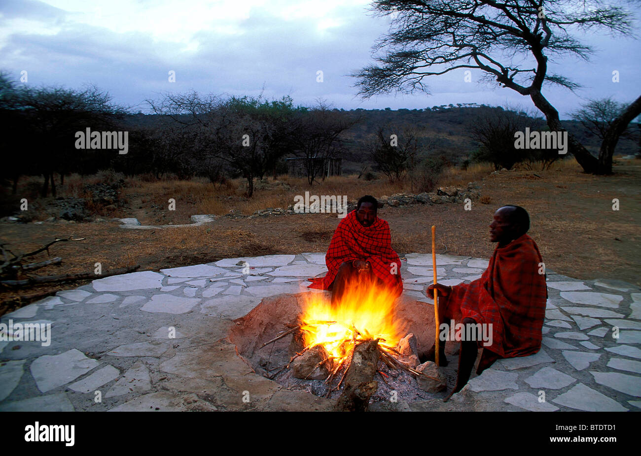 Zwei schwarze Masaai Männer gekleidet in ihren traditionellen roten Shukas sitzen um ein Feuer Stockfoto