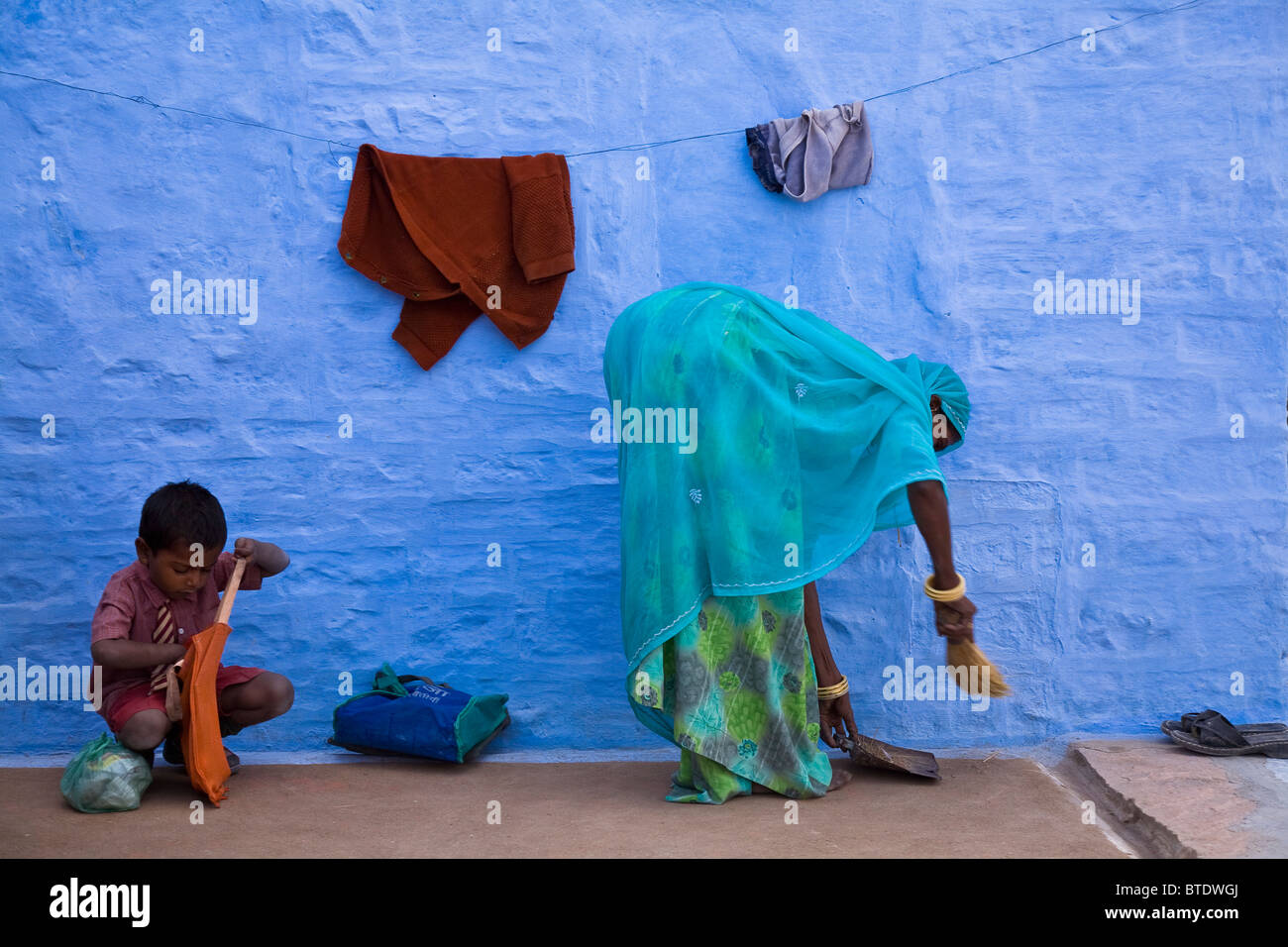 Eine indische Frau fegt ihr Hinterhof, als ihr Kind bereitet sich für den Schulunterricht, Salawas, Rajasthan, Indien zu verlassen. Stockfoto