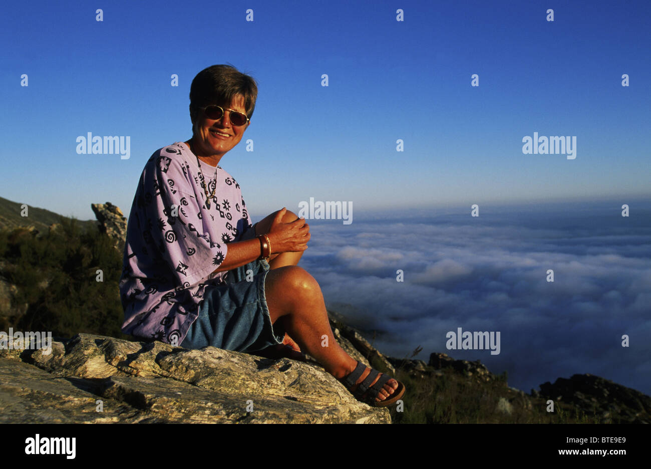 Eine lächelnde Dame sitzt auf einem Felsen über den Wolken auf dem Swartberg Pass (Modell freigegeben) Stockfoto