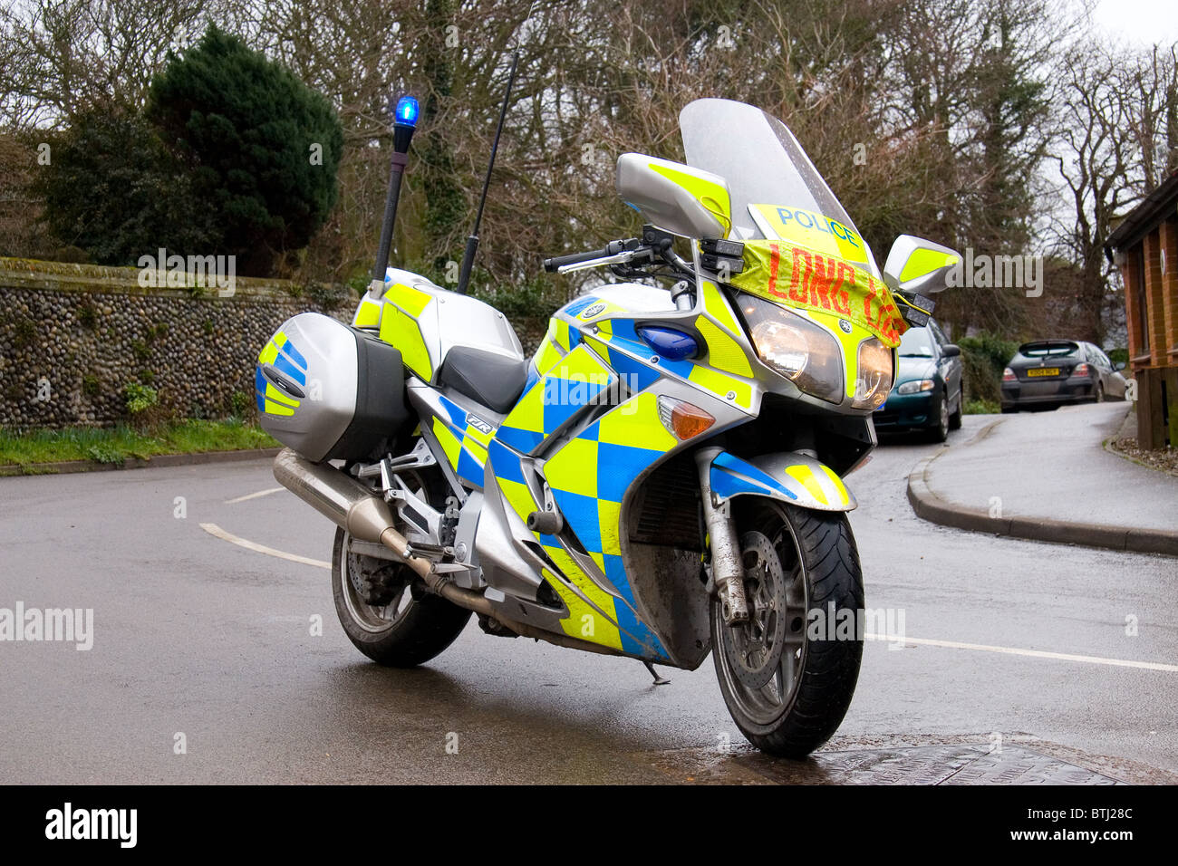 Britisches Polizeimotorrad mit blauen Notlichtern und „Long Load“-Meldung über ungewöhnliche Last, geparkt auf einer nassen Dorfstraße in Norfolk, England, Großbritannien. Stockfoto