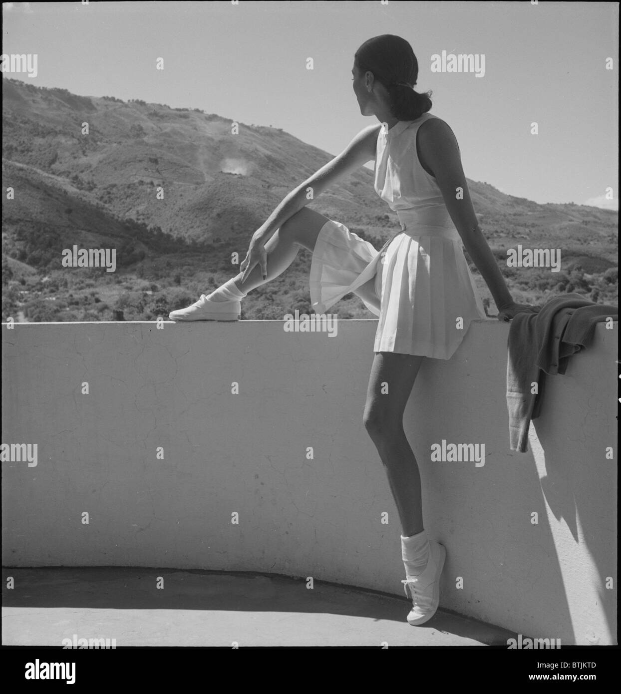 Frau trägt Tennis-Outfit, auf Wand, mit einem Bein auf Wand, sitzend mit Blick auf die Berge hinter ihr, Foto von Toni Frissell, 1947. Stockfoto