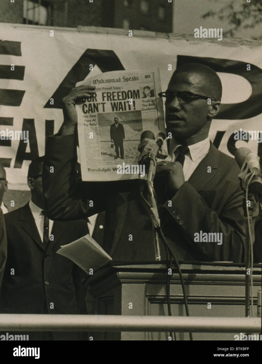 Malcolm X, hochhalten Zeitung mit der Schlagzeile "Unsere Freiheit can't Wait," stehend hinter Podium mit Mikrofonen, die Adressierung einer Menschenmenge auf Black Muslim rally in New York. 1963. Stockfoto