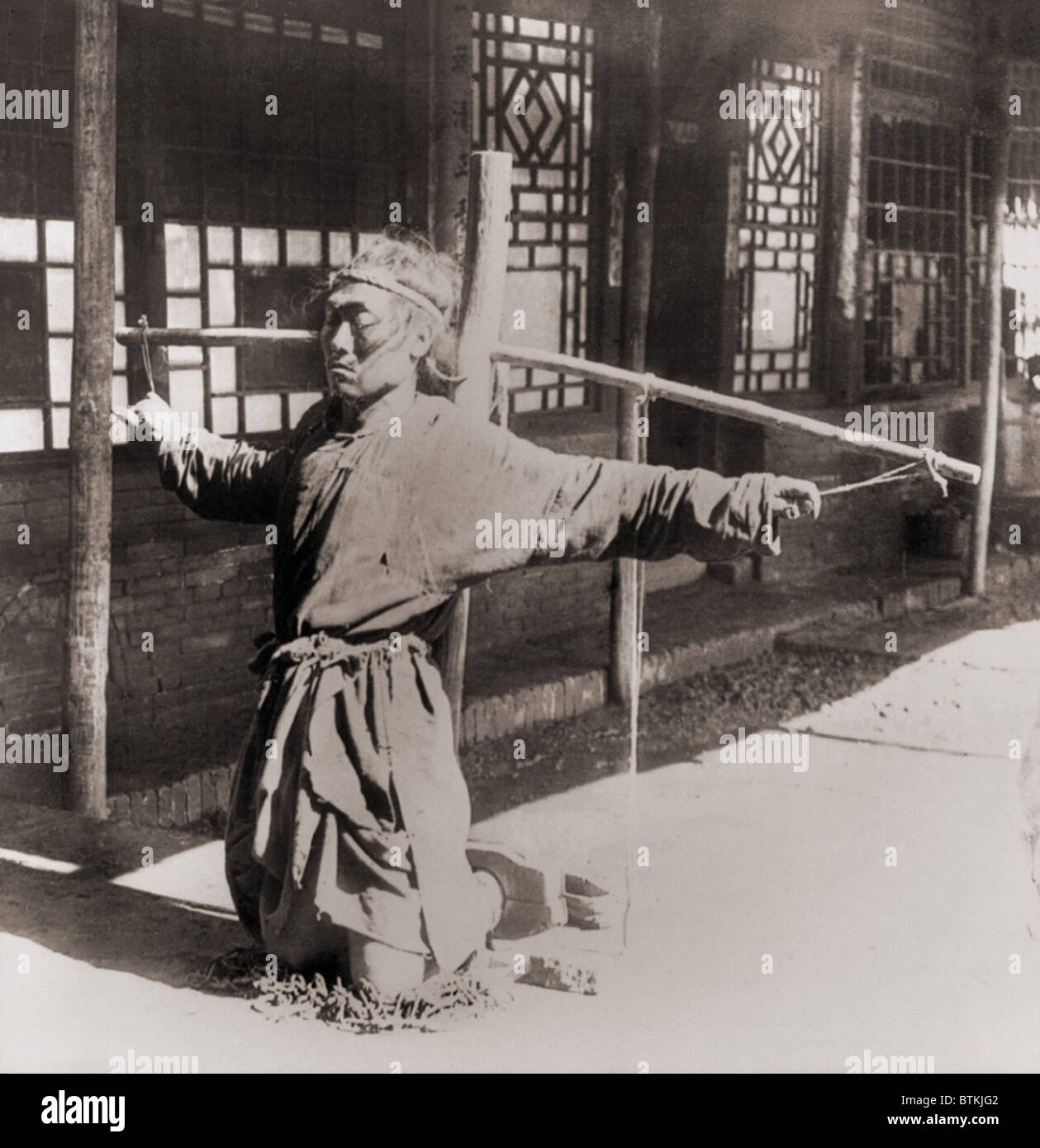 Chinesische Gefangene kniend auf Ketten, mit den Knöcheln im Bestände, während seine Arme durch die Daumen an einen Pfahl unterstützt seine Arme gebunden sind. Moukden, China. Ca.. 1900. Stockfoto