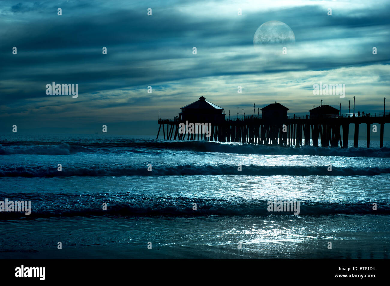 Ein schöner Abend am Strand entlang zeigt einen Pier in einem niedrigen Mond mit Spiegelungen im Wasser und ein stimmungsvoller Himmel. Stockfoto