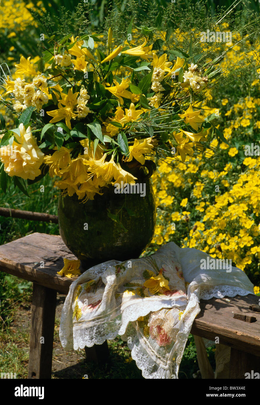 Stillleben von gelben Freesien und Lilien in Vase auf Gartenbank gegen gelb blühender Strauch Stockfoto