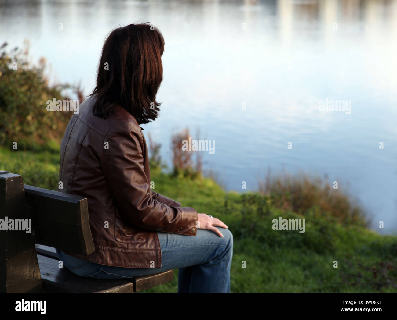 Rückansicht einer Frau tief in Gedanken mit Blick auf einen See, auf einer Parkbank sitzen. Stockfoto