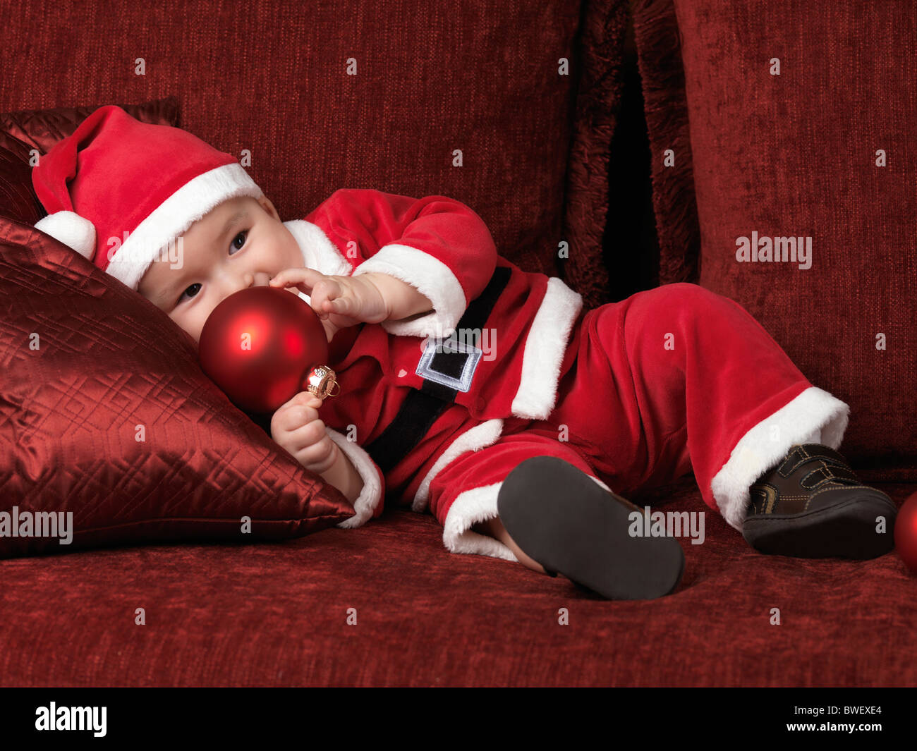 Lizenz erhältlich bei MaximImages.com – ein sechs Monate alter Junge im Weihnachtsmann-Kostüm, der eine rote Kugel in der Hand hält Stockfoto