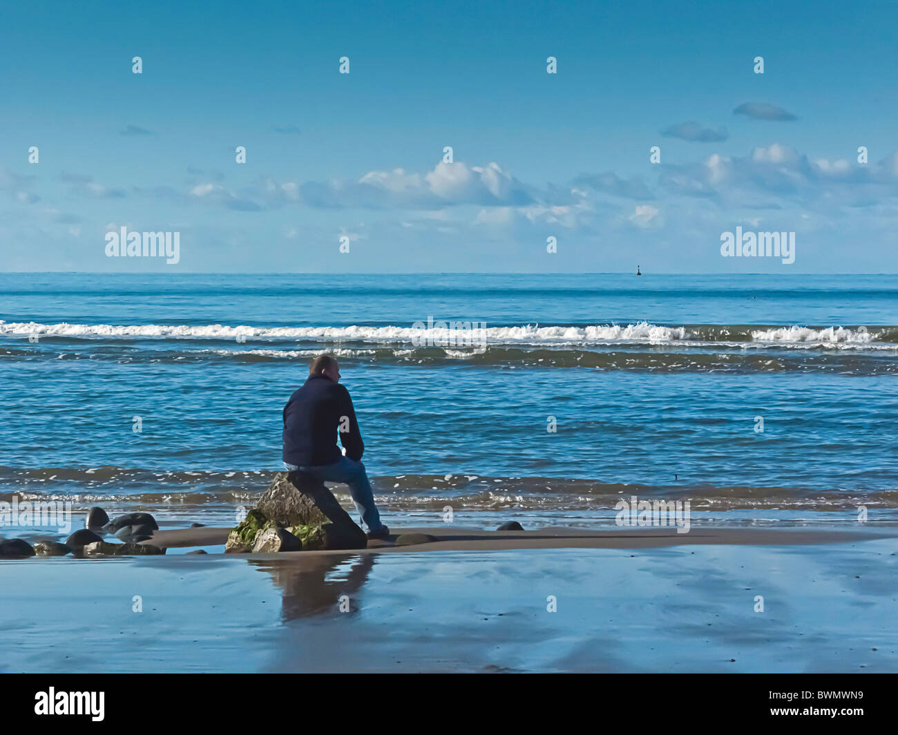 Mann allein zu sitzen, auf Felsen, Blick auf das Meer, Sandstrand, Wellen brechen, blauer Himmel, Horizont Stockfoto