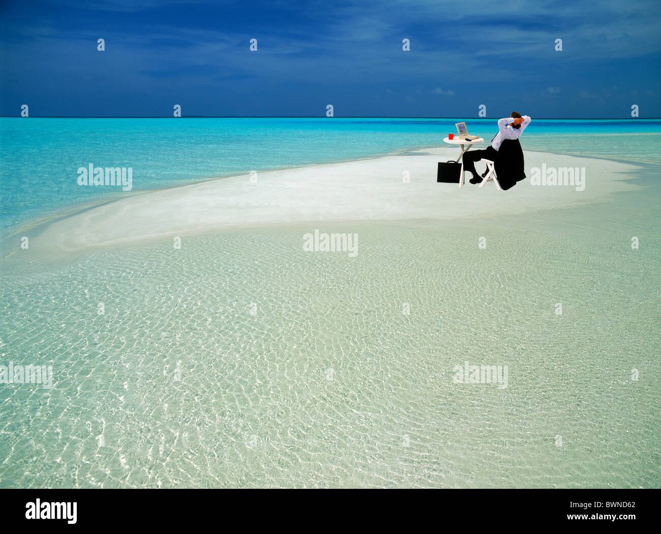 Geschäftsmann im Anzug mit Aktentasche sitzt im Stuhl am Tisch mit Laptop und Kaffee Tasse auf abgelegenen Sandbank in reine Einsamkeit Stockfoto