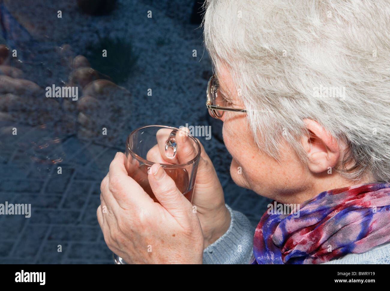 Nachdenklich, ältere Frau mit einem warmen Getränk mit Blick durch ein Fenster in einer kalten, dunklen Nacht mit Schnee im Winter. Großbritannien Großbritannien Stockfoto