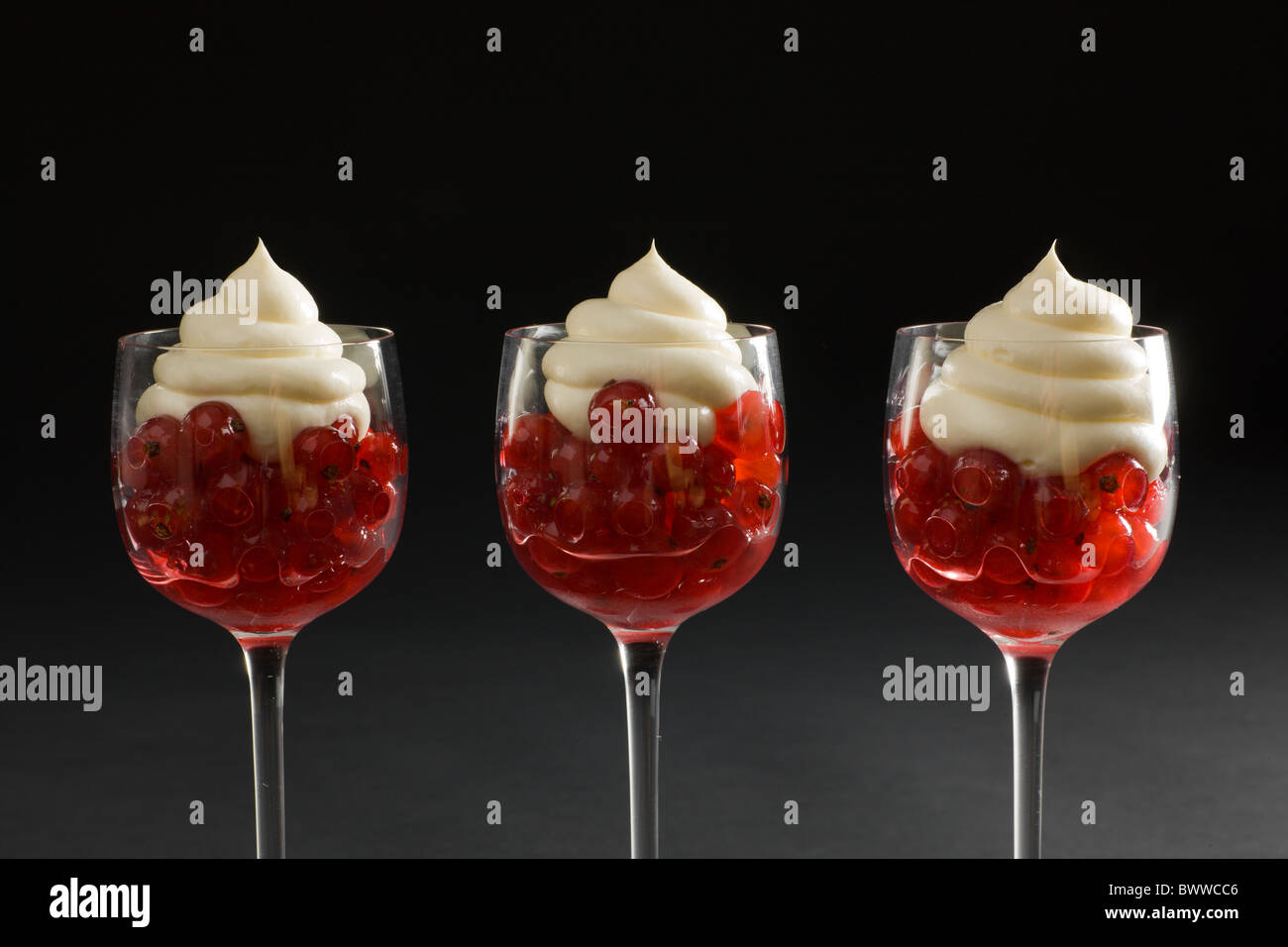 Rote Johannisbeeren in einem würzigen Sirup in eleganten Stiel Gläser, gekrönt mit einer cremigen Wirbeln auf einem dunklen Hintergrund. Stockfoto