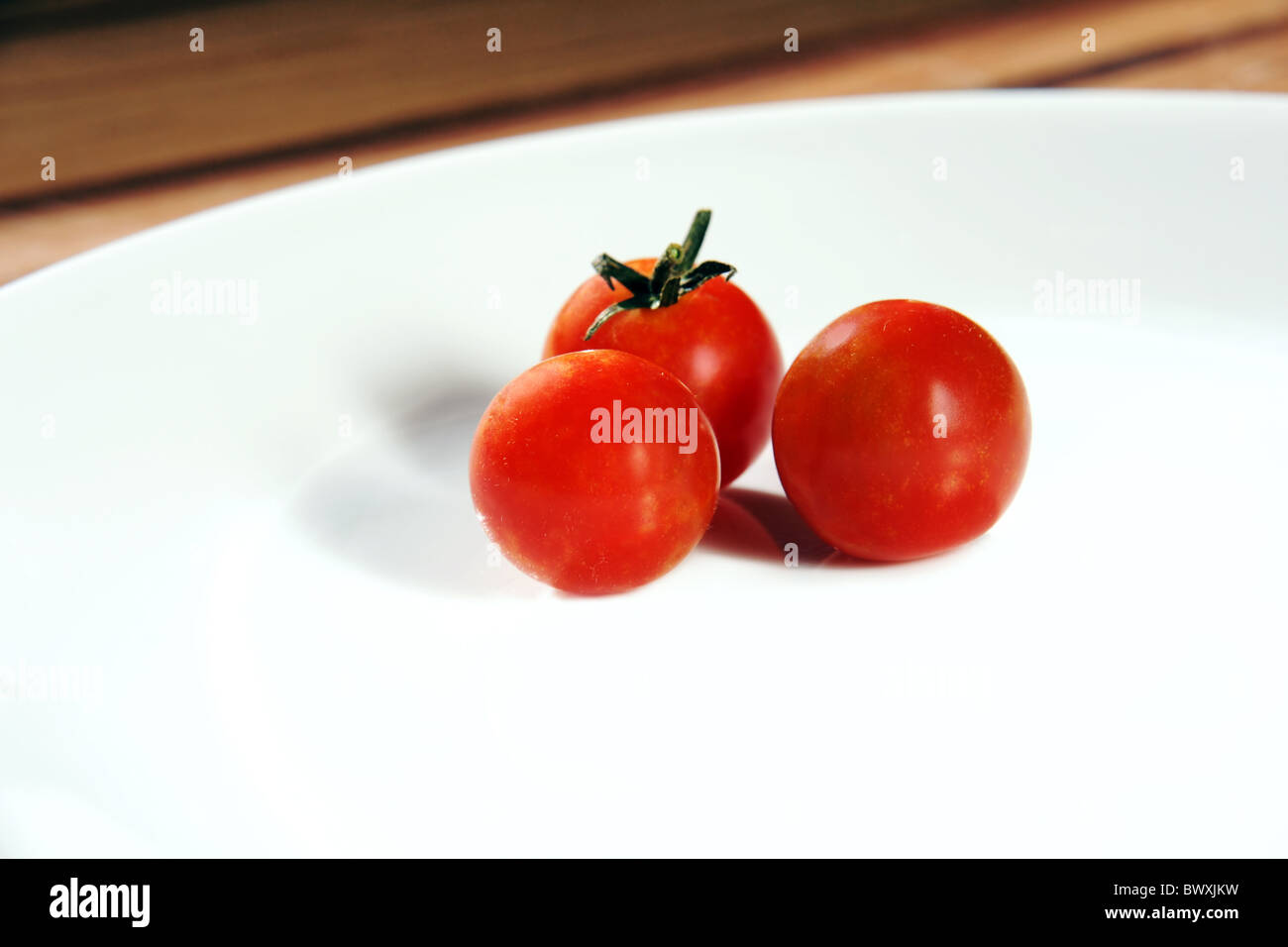 Drei helle rote, Reife Cherry Tomaten auf einem knackigen weißen Porzellanteller platziert auf einem Holztisch. Stockfoto