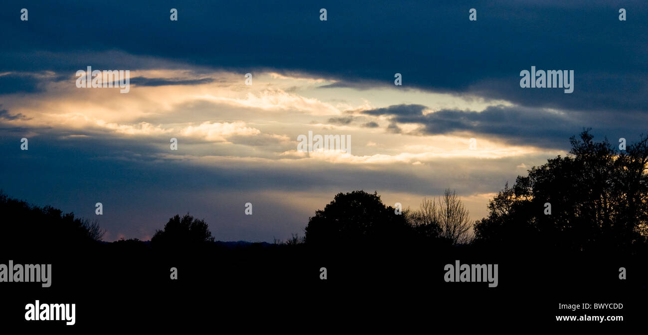 Stimmungsvolle Landschaft Skyscape mit Öffnung in den Wolken zeigt Abend Sonnenlicht durch und Bäume im Sillouette unten Stockfoto