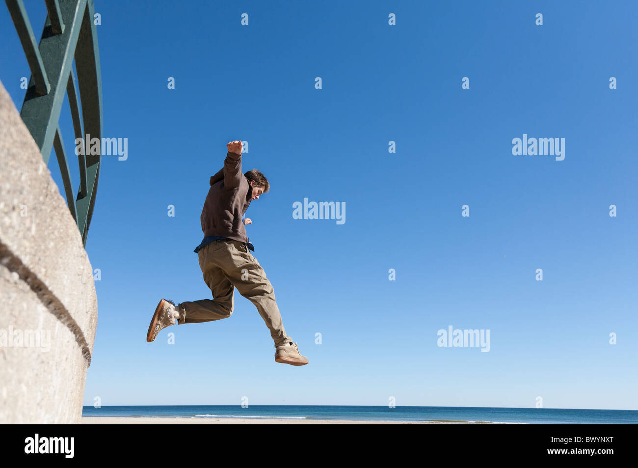 Ein junger weißer Mann springt aus einer Wand. Lake Michigan ist im Hintergrund sichtbar. Es ist ein sonniger Tag mit einem tiefblauen Himmel. Stockfoto