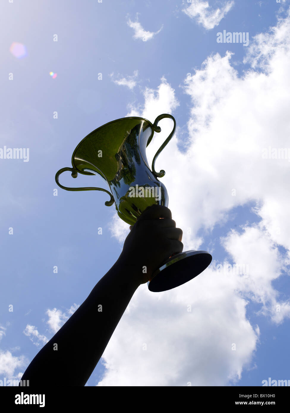 Eine siegreiche Person hält einen großen, glänzenden Trophäenbecher hoch, der sich vor einem leuchtend blauen Himmel mit ein paar Wolken bildet. Stockfoto