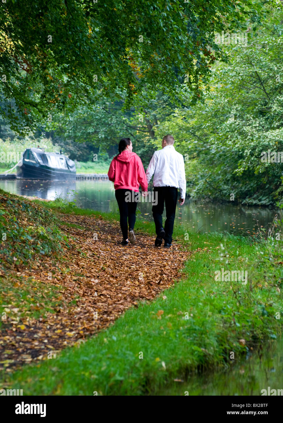 Paar hand in hand gehen entlang Kanal Leinpfad von Kamera mit Kanal und Narrowboat oder Schiff festgemacht vor Ihnen Stockfoto