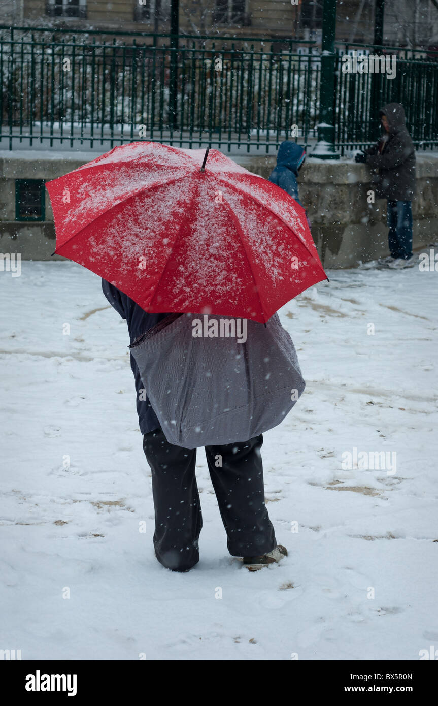 Eine Mutter unter einem roten Regenschirm Uhren ihre Kinder spielen im Schnee in einem Park in Paris. Stockfoto