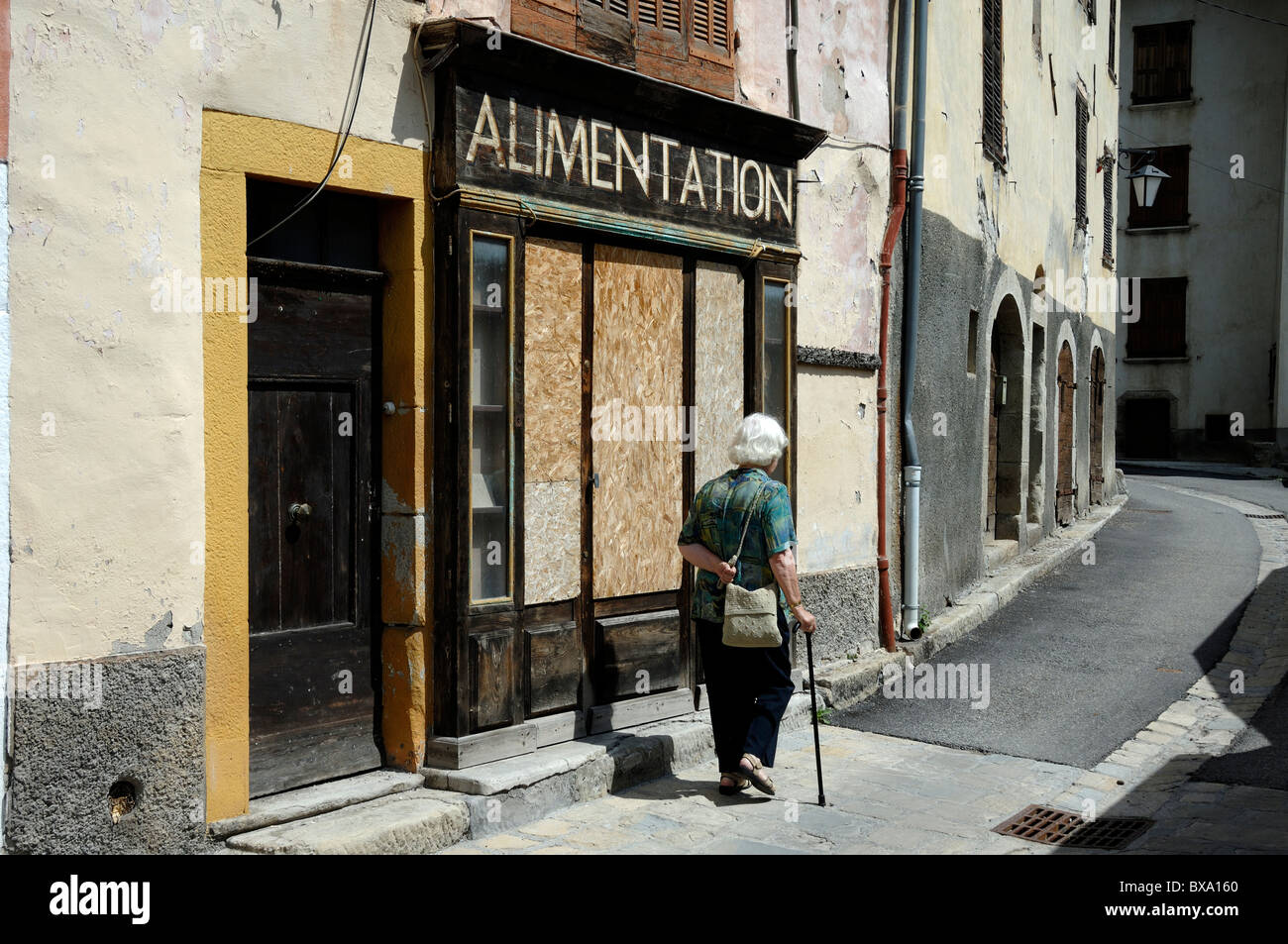 Die alte Frau mit Spazierstock geht an einem vernagelten und geschlossenen Dorfladen oder General Store, Alimentation, Beauvezer, Alpes-de-Haute-Provence, Frankreich vorbei Stockfoto
