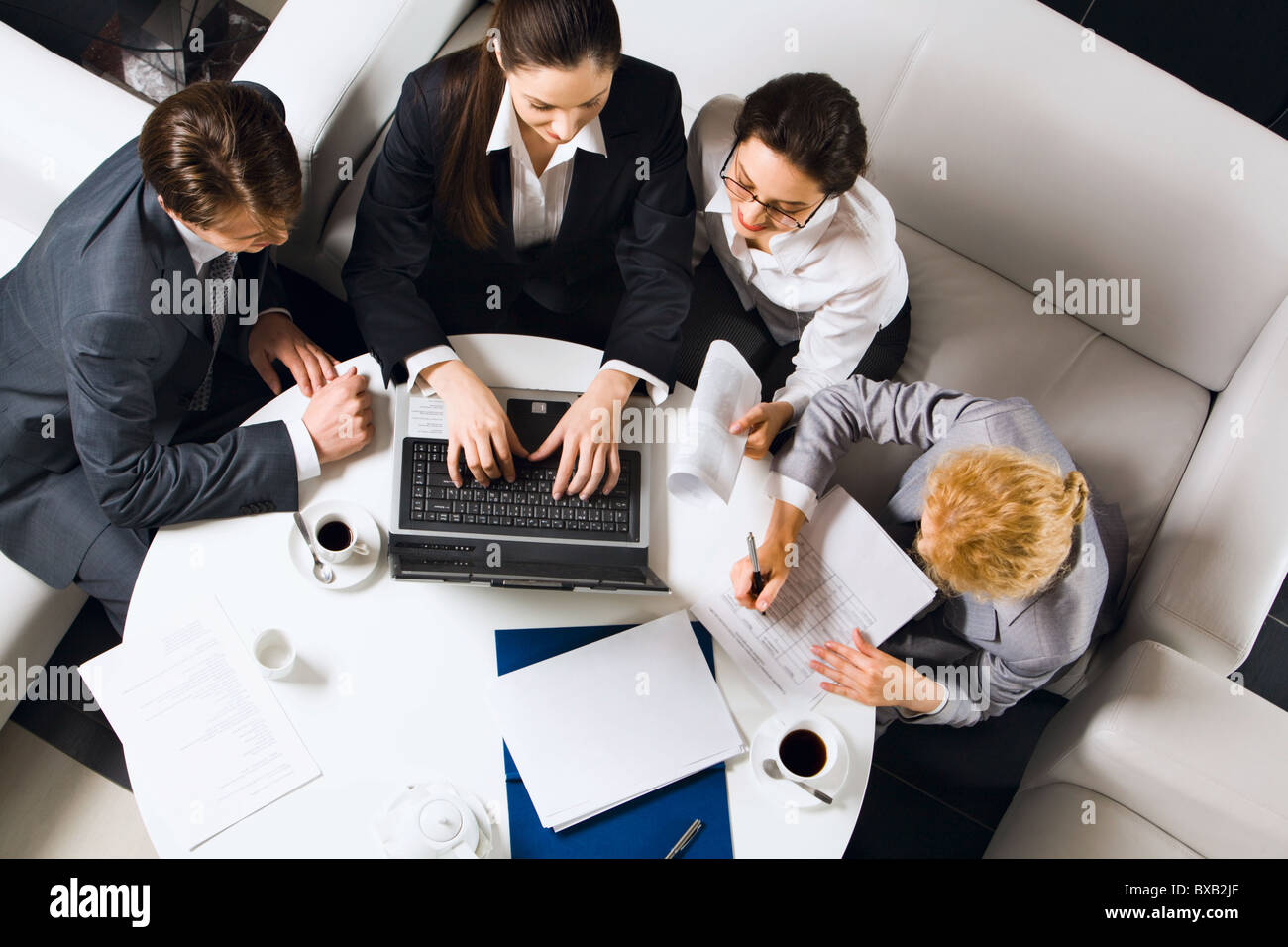 Drei Unternehmerinnen und Unternehmer sitzen auf den weißen Sofas am runden Tisch mit geöffneten Laptop, Dokumente und Tassen von co Stockfoto