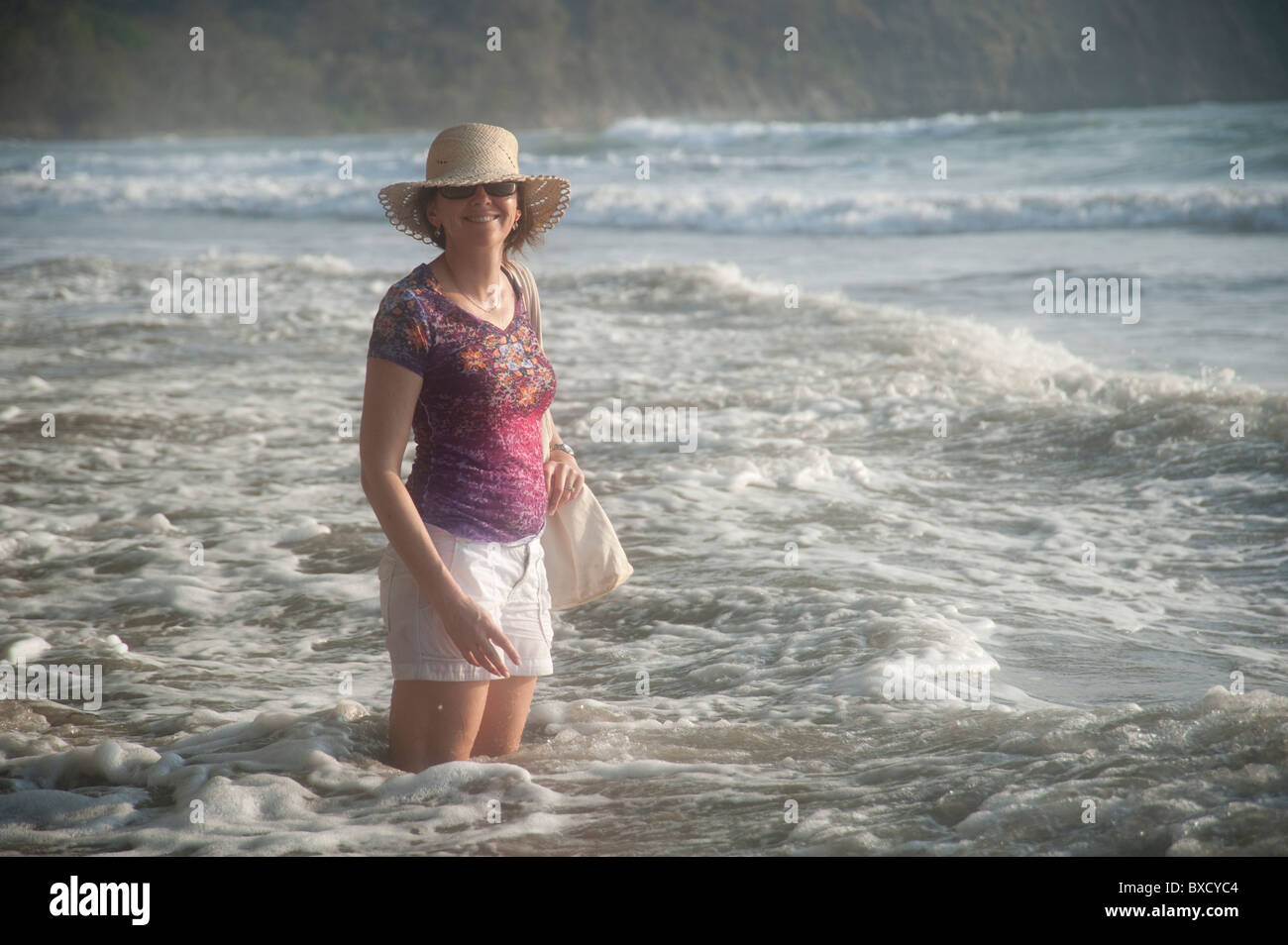 Frau mit Hut lächelnd ansehen in der Meeresbrandung auf Kniehöhe Stockfoto