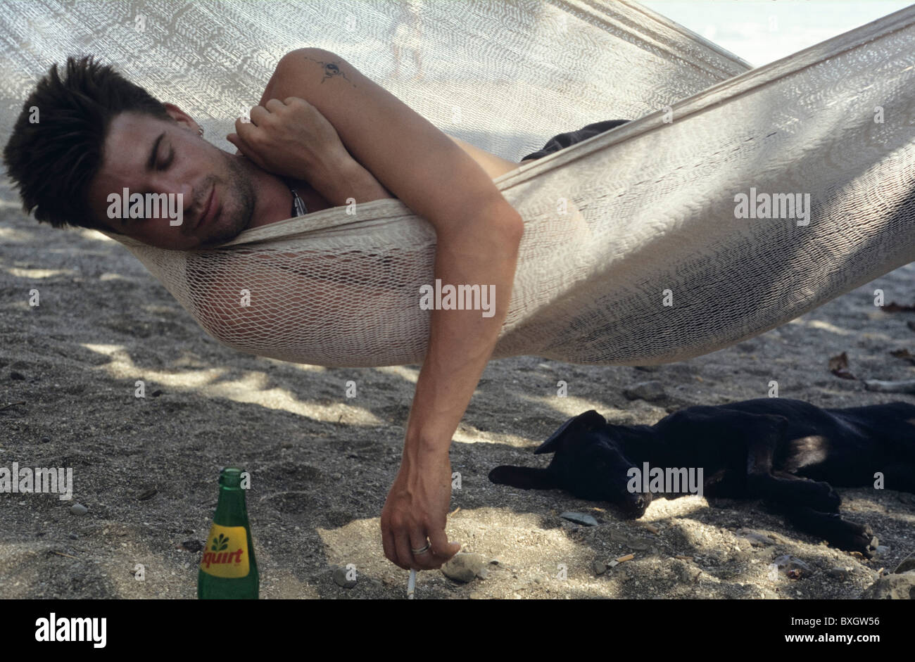 Costa Rica, junger Mann in Montezuma dösen in Hängematte mit Zigarette in der hand, schwarze kleine Hund schlafen im Schatten, alkoholfreies Getränk Stockfoto