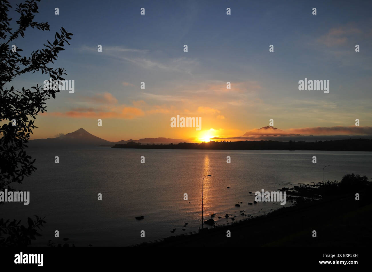 Llanquihue See Blick von Puerto Varas, der eine gelbe Sonne durch Wolken zwischen Osorno und Calbuco Vulkane, Chile Stockfoto