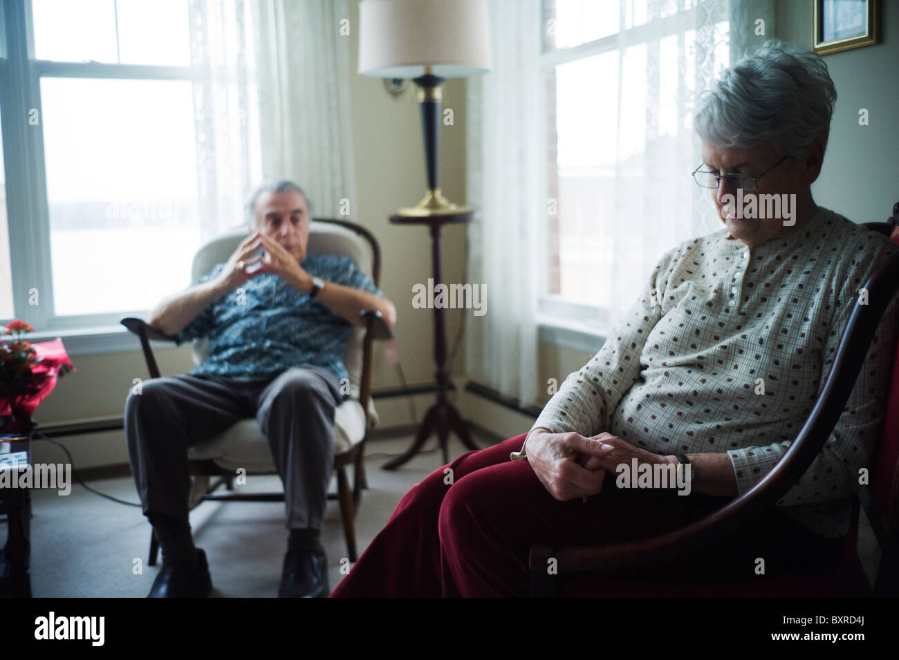 Alte Frau sitzt im Vordergrund mit den Händen umklammert, während Alter Mann aus Hintergrund schaut Stockfoto