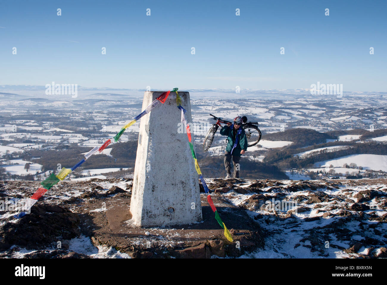 Mann trägt ein Mountainbike im Schnee auf den Hügeln von Malvern, Worcestershire, mit Gebetsfahnen auf der Triangulation Punkt Stockfoto