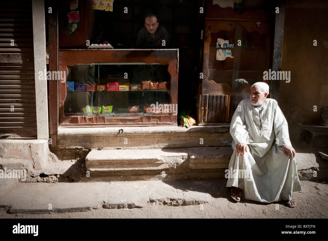 Mann sitzt auf dem Bürgersteig vor kleinen Laden als Shop Keeper schaut aus Zähler bei einem örtlichen Straßenmarkt, Luxor, Ägypten, Afrika Stockfoto