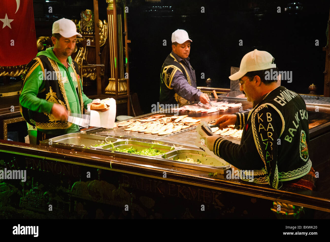 Balik Ekmek Fish Sandwich Vendor Istanbul Turkey // ISTANBUL, Türkei — Ein Händler grillt frischen Fisch auf einem traditionellen balıkçı teknesi (Fischerboot), das im Goldenen Horn nahe Eminönü und der Galata-Brücke vor Anker liegt. Diese schwimmenden Küchen servieren Balık ekmek, Istanbuls ikonisches Fisch-Sandwich mit gegrillter Makrele, serviert in Weißbrot mit Zwiebeln und Salat. Die kunstvoll verzierten Boote, oft mit osmanischen Motiven mit Goldverzierungen und halbmondförmigen Finialen versehen, liegen in der Nähe der Fährhafen Eminönü fest. Diese jahrhundertealte Tradition der Fischverkäufer am Wasser ist nach wie vor so weit Stockfoto
