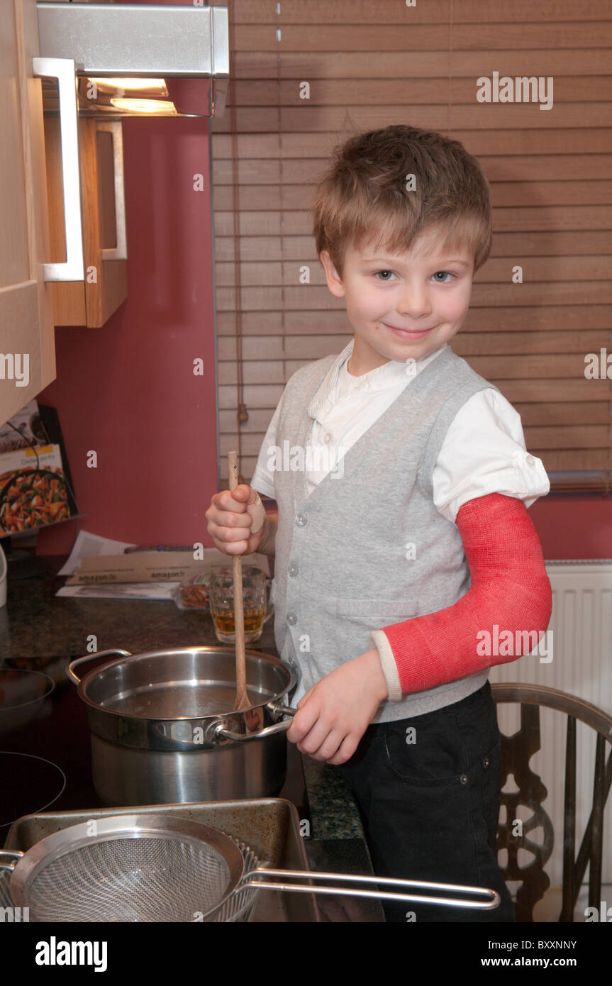 vier-jähriger Junge mit einem gebrochenen Arm Kochen machende Kuchen auf einem Stuhl neben dem Kochplatten und Backofen Stockfoto