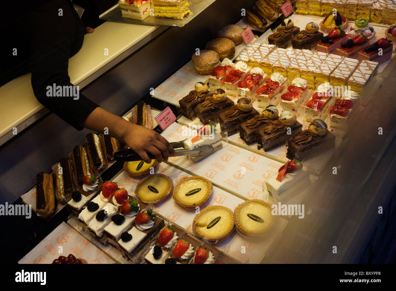 Eine anonyme Hand drückt ein paar Zangen, Sahne-Torte von einem Display in Patisserie Valerie in London zu wählen. Stockfoto