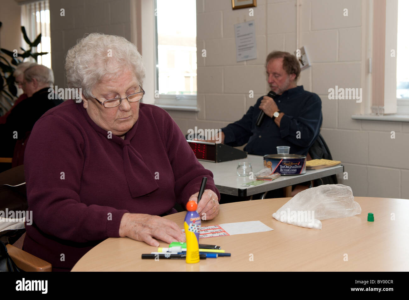 Ältere Dame saß am Tisch spielen Bingo in einem Tageszentrum für die 50er Jahre mit freiwilligen Bingoanrufer im Hintergrund. Stockfoto