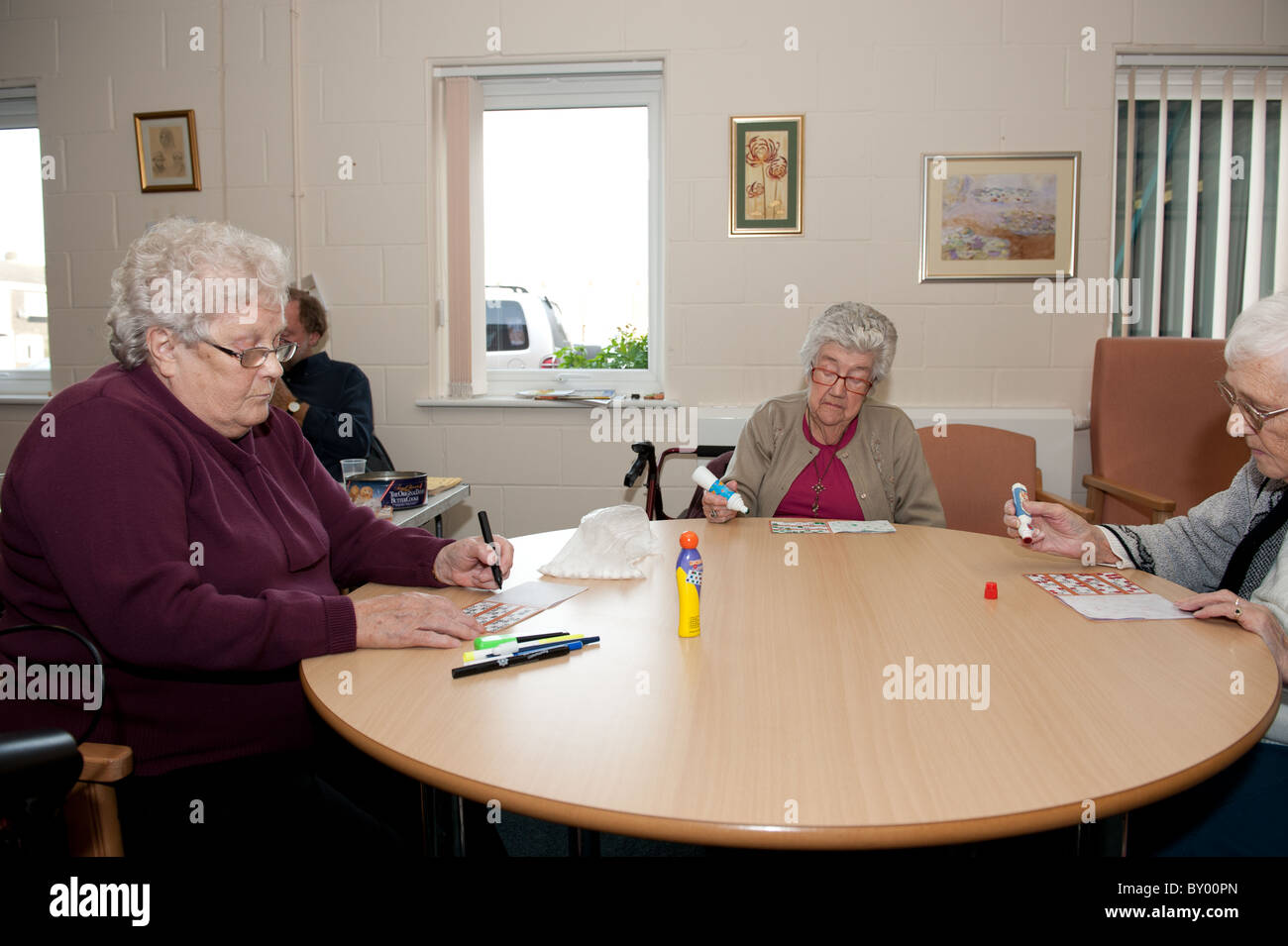 Drei ältere Freunde sitzen an Tisch im Tageszentrum Bingo zusammen zu spielen. Männliche Bingoanrufer im Hintergrund. Stockfoto