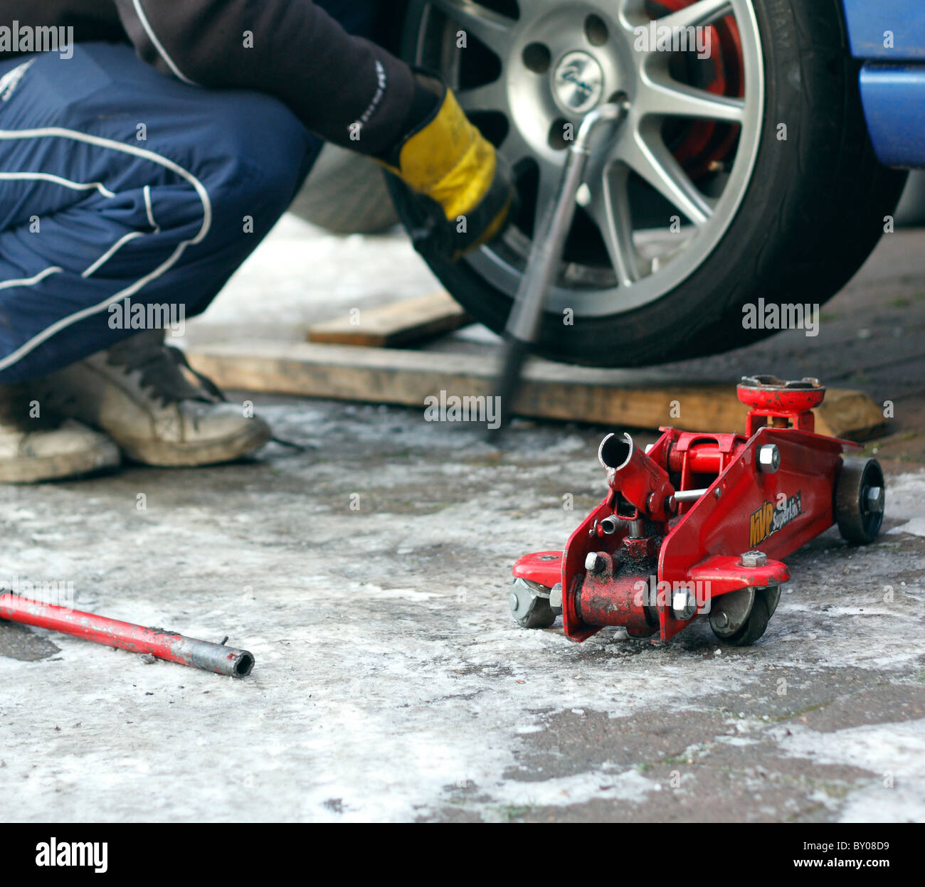 Eine rote hydraulische Rangierwagenheber neben einem Auto, während ein Mechaniker ist die Radmuttern mit einem Schraubenschlüssel auf das Rad abnehmen abschrauben. Stockfoto