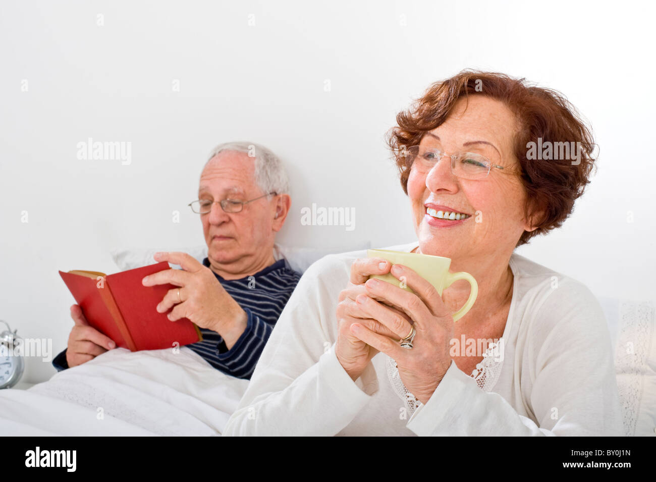 Älteres Paar im Bett Mann liest und Frau, die eine Tasse Kaffee Stockfoto