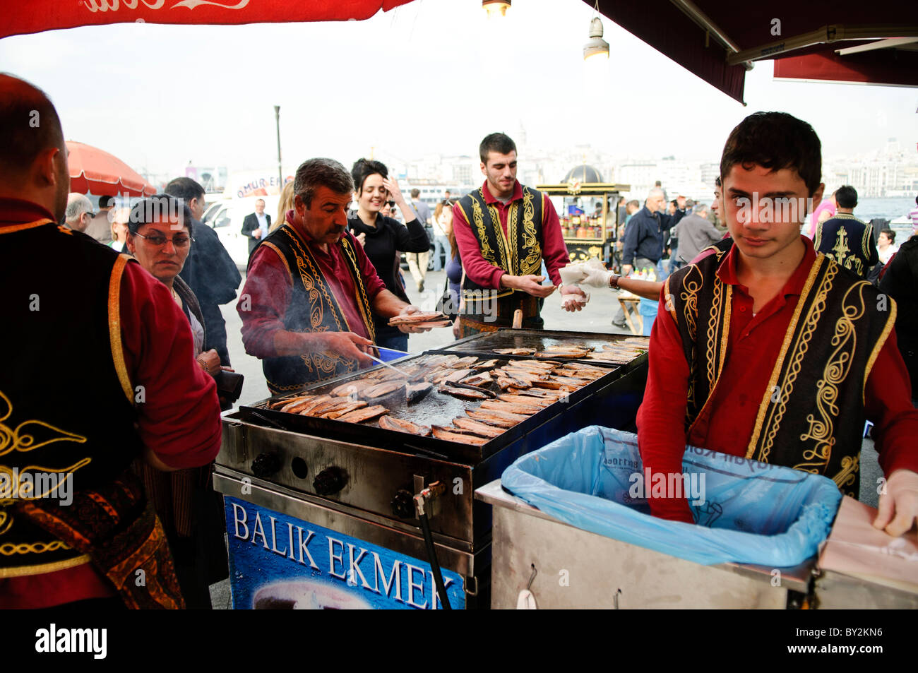 Balik Ekmek Fish Sandwich Street Food Istanbul Turkey // ISTANBUL, Türkei – Ein Straßenverkäufer grillt frischen Fisch am Ufer von Eminonu in der Nähe der Galata-Brücke und bereitet das traditionelle balik ekmek (Fischsandwich) zu. Der balik ekmek, der typischerweise aus gegrillter Makrele in Weißbrot mit Zwiebeln und Salat zubereitet wird, gehört zu den beliebtesten Street Food-Gerichten Istanbuls. Fischverkäufer am Wasser bedienen sich seit Generationen von schwimmenden Booten und Ständen entlang des Goldenen Horns. Der Stadtteil Eminonu dient als wichtiger Fährhafen und Handelsknotenpunkt zwischen Istanbuls Europa und Asien Stockfoto