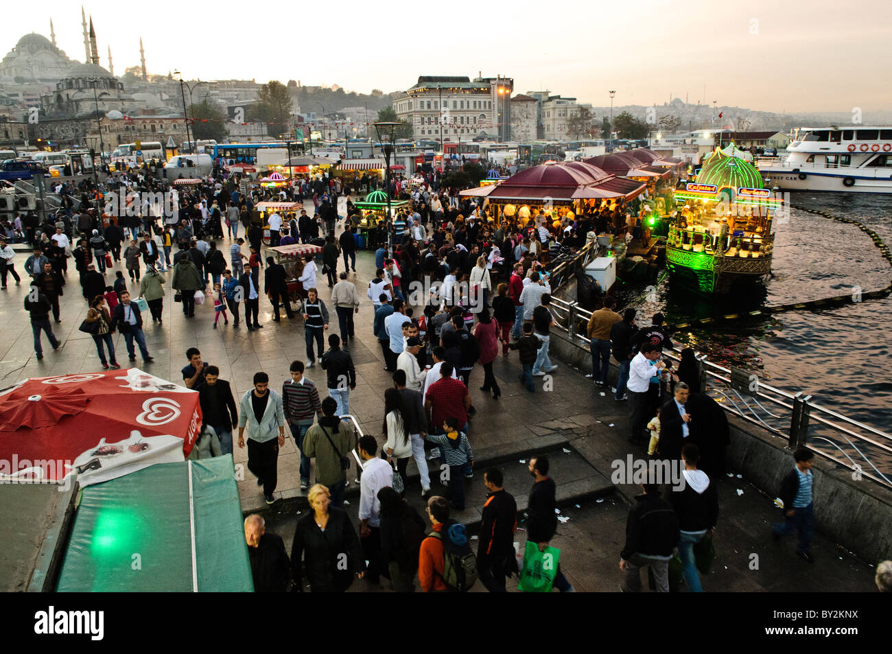 Balik Ekmek Fisch-Sandwiches Eminonu Waterfront Istanbul // ISTANBUL, Türkei - beleuchtete Fischerboote servieren traditionelle balik Ekmek (Fisch-Sandwiches) entlang der belebten Eminonu-Uferpromenade am Fuße der Galata-Brücke. Die abendliche Szene fängt die lebendige Esskultur am Wasser mit historischen Moscheen ein, die sich vor dem dunklen Himmel verbergen. Die lokalen Händler führen eine jahrhundertealte Tradition fort, frischen gegrillten Fisch von ihren Booten aus zu servieren. Stockfoto