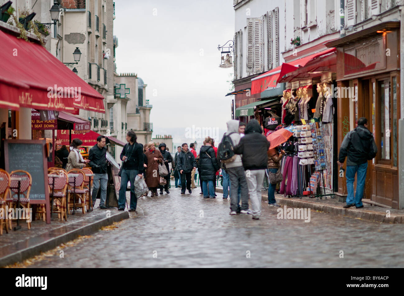 Montmartre Cobblestone Street Cafe Szene mit Menschen Paris France // PARIS, Frankreich – Eine kopfsteingepflasterte Straße in Montmartre bietet traditionelle Cafés mit roten Markisen und historischen Gebäuden, die von Fußgängern bevölkert werden. Dieses berühmte Viertel im 18. Arrondissement von Paris ist bekannt für sein künstlerisches Erbe und seine charmanten, verwinkelten Straßen. Montmartre war historisch ein Drehkreuz für Künstler wie Pablo Picasso und Vincent van Gogh, was zu seinem einzigartigen Bohemiencharakter beitrug. Heute zieht die einzigartige Atmosphäre und die malerische Umgebung zahlreiche Besucher und Kunstliebhaber an Stockfoto